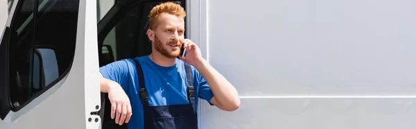 Панорамний знімок завантажувача в комбінезоні, що розмовляє на смартфоні біля вантажівки на відкритому повітрі — стокове фото