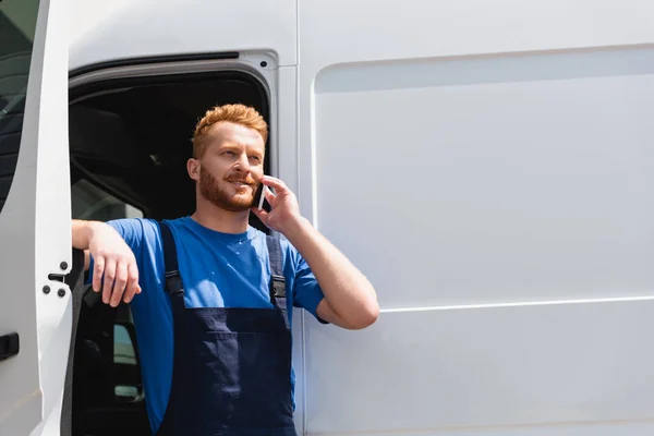 Погрузчик в комбинезоне разговаривает по смартфону, стоя рядом с грузовиком с открытой дверью — стоковое фото