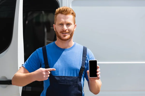 Loader in overalls pointing at smartphone with blank screen near truck outdoors — Stock Photo