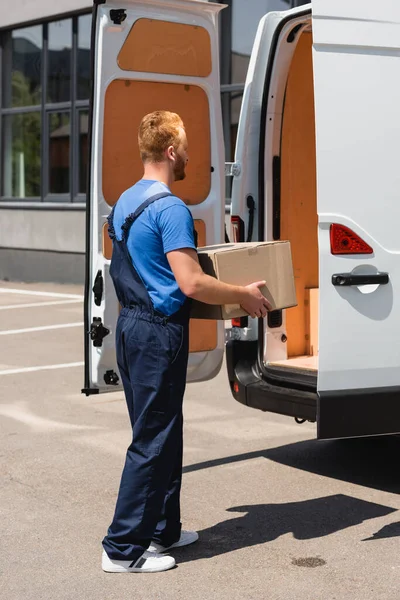 Loader in overalls holding carton box near truck with open doors on urban street — Stock Photo