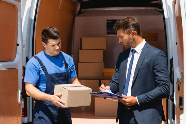 Businessman writing on clipboard near loader holding carton box beside truck outdoors — Stock Photo