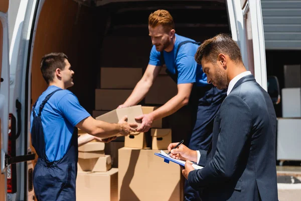 Focus selettivo di uomo d'affari che scrive sugli appunti mentre i traslocatori tengono scatola di cartone in camion all'aperto — Foto stock