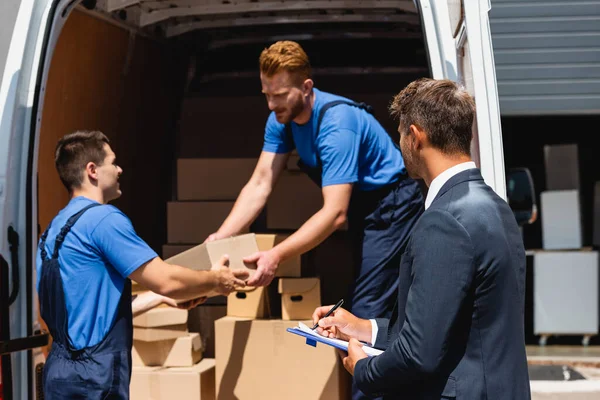 Selective focus of businessman holding pen and clipboard while movers unloading packages from truck outdoors — Stock Photo