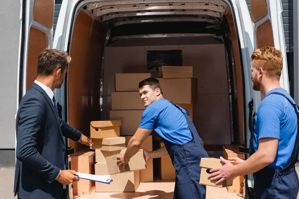 Businessman with clipboard pointing with hand while loader holding cardboard box in truck outdoors — Stock Photo