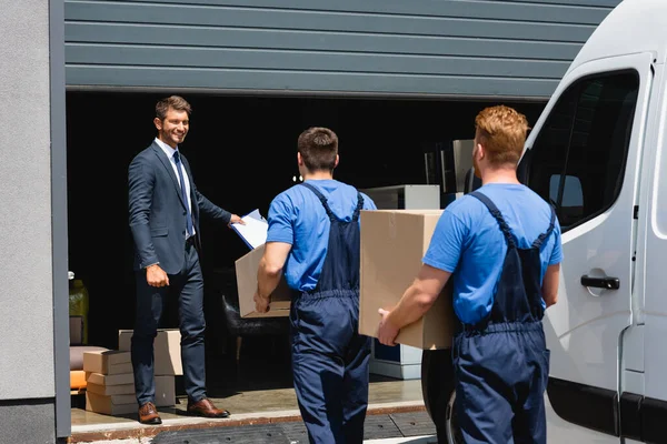 Selective focus of businessman holding clipboard while movers carrying packages near truck and warehouse outdoors — Stock Photo