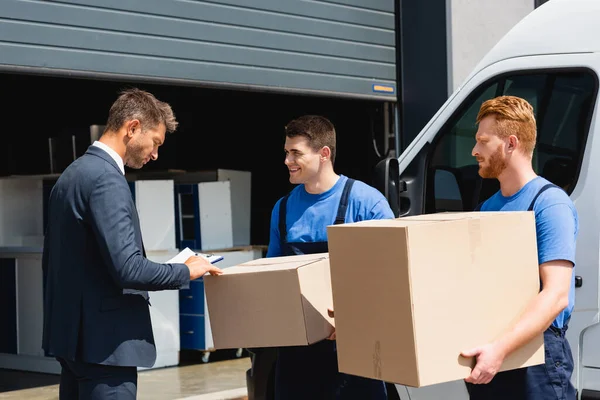 Déménageurs avec boîtes en carton debout près de l'homme d'affaires avec presse-papiers et camion sur la rue urbaine — Photo de stock