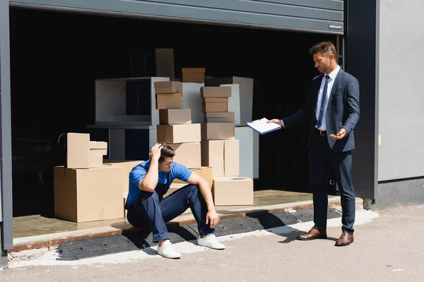 Businessman in suit holding clipboard and pointing with hand near sad loader in warehouse — Stock Photo