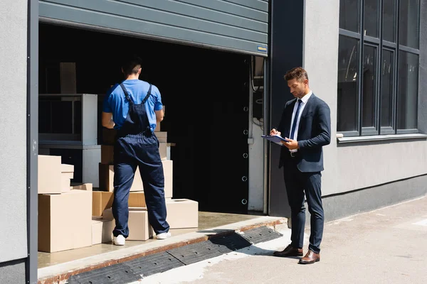 Empresário escrevendo na área de transferência ao lado do carregador que trabalha no armazém na rua urbana — Fotografia de Stock