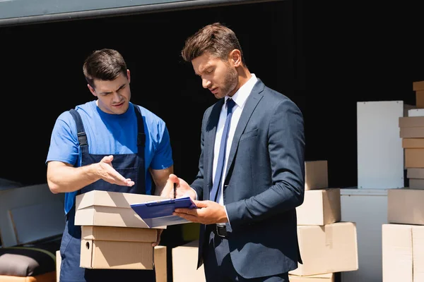 Cargador con cajas de cartón apuntando con la mano cerca de hombre de negocios con portapapeles y almacén en el fondo - foto de stock