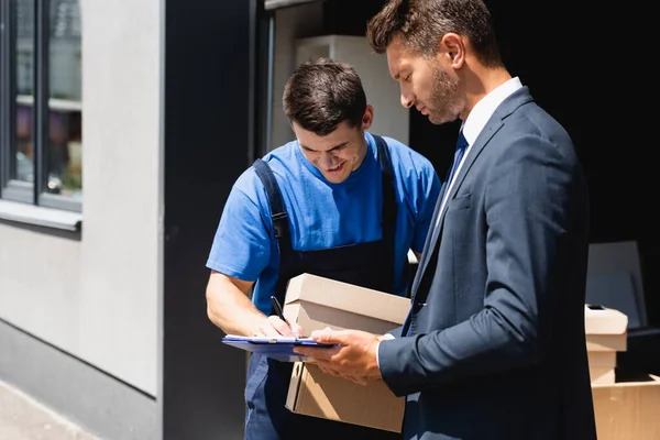 Loader with cardboard boxes writing on clipboard near businessman on urban street — Stock Photo