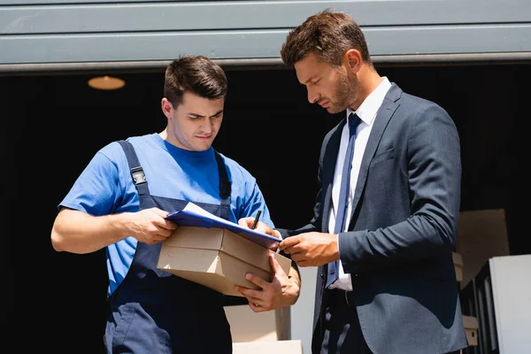 Loader holding carton box while businessman in suit writing on clipboard with warehouse at background — Stock Photo