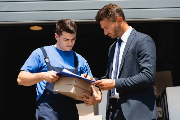 Caricatore contenente scatola di cartone mentre l'uomo d'affari in giacca e cravatta scrive sugli appunti con magazzino sullo sfondo — Foto stock