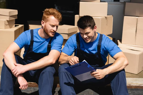 Movers looking at clipboard while sitting near cardboard boxes in warehouse — Stock Photo