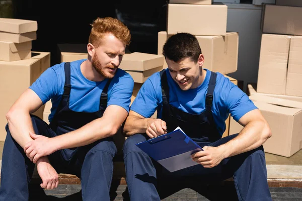 Umzugsteilnehmer betrachten Klemmbrett, während sie in der Nähe von Kartons im Lager sitzen — Stockfoto