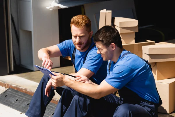 Movers in overalls looking at clipboard near carton boxes in warehouse — Stock Photo