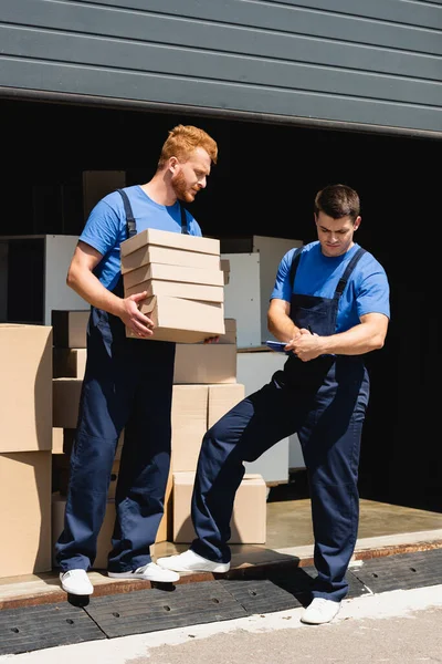 Movers working with cardboard boxes and clipboard near warehouse outdoors — Stock Photo