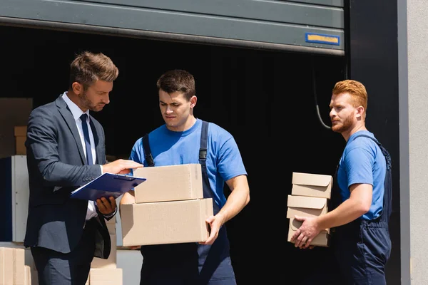 Businessman with clipboard checking carton boxes near loaders and warehouse outdoors — Stock Photo