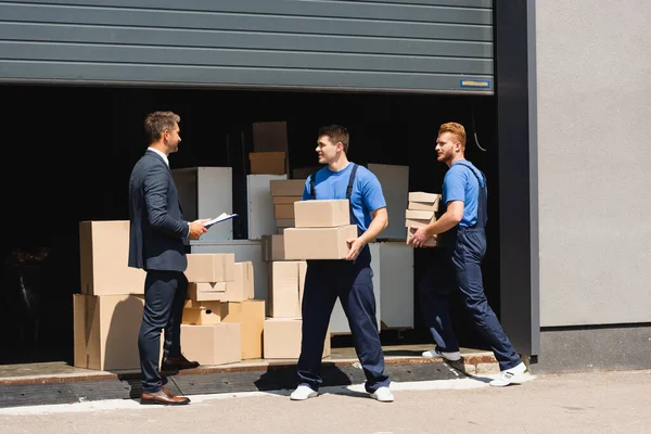 Homem de negócios em terno segurando prancheta perto de movers com pacotes e armazém ao ar livre — Fotografia de Stock