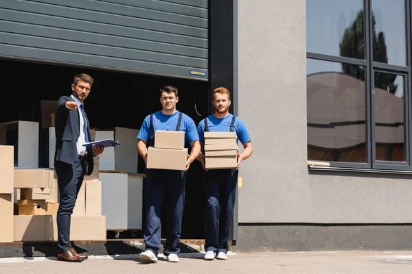 Businessman with clipboard pointing with hand near movers with cardboard boxes and warehouse outdoors — Stock Photo