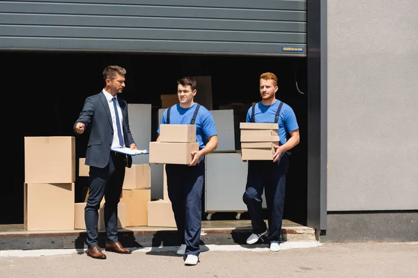 Man in formal wear holding clipboard and pointing with hand near movers with carton boxes and warehouse — Stock Photo