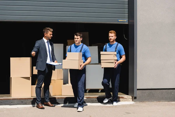 Hombre en desgaste formal sujetando portapapeles y apuntando con la mano cerca de los transportistas con cajas de cartón y almacén - foto de stock