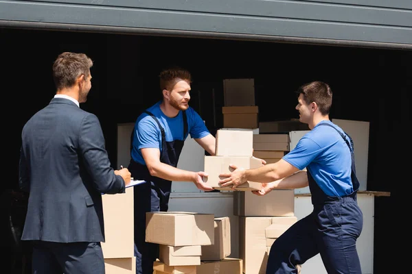 Déménageurs travaillant avec des boîtes en carton dans l'entrepôt tandis que l'homme d'affaires écrit sur le presse-papiers à l'extérieur — Photo de stock