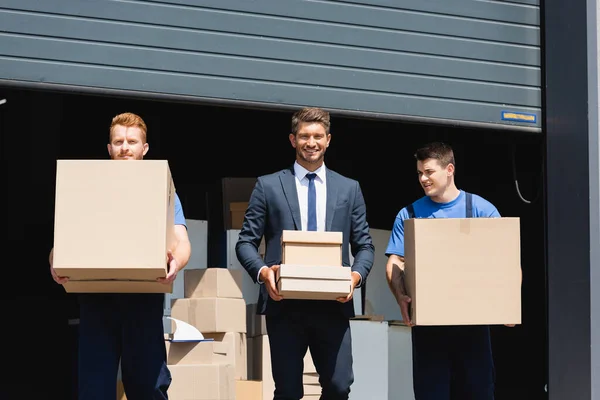 Businessman holding cardboard boxes near loaders with warehouse on background — Stock Photo