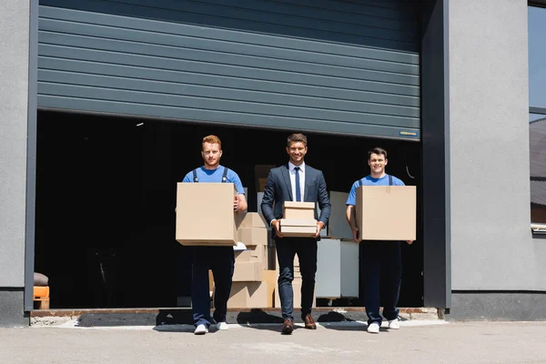 Бизнесмен в костюме и грузчики смотрят в камеру, держа коробки возле склада на городской улице — стоковое фото