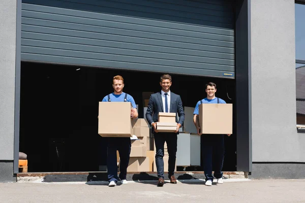 Homme d'affaires en costume et déménageurs regardant la caméra tout en tenant des boîtes en carton près de l'entrepôt dans la rue urbaine — Photo de stock