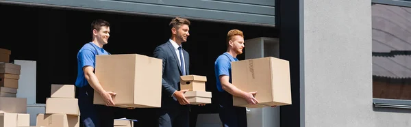 Horizontal crop of loaders and businessman walking with cardboard boxes near warehouse on urban street — Stock Photo