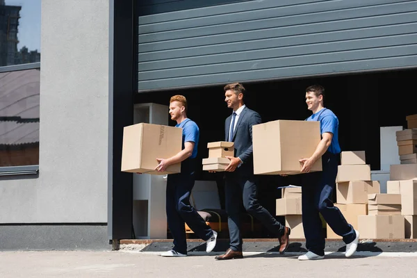 Empresário segurando caixas de papelão enquanto caminha com movers na rua urbana — Fotografia de Stock