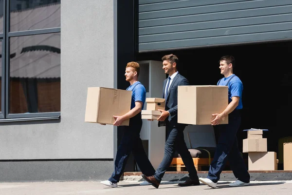 Movers carregando caixas de papelão enquanto caminhava perto de empresário na rua urbana — Fotografia de Stock