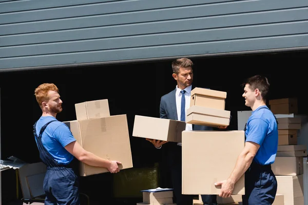 Movers com caixas de papelão em pé perto de empresário em terno e armazém ao ar livre — Fotografia de Stock