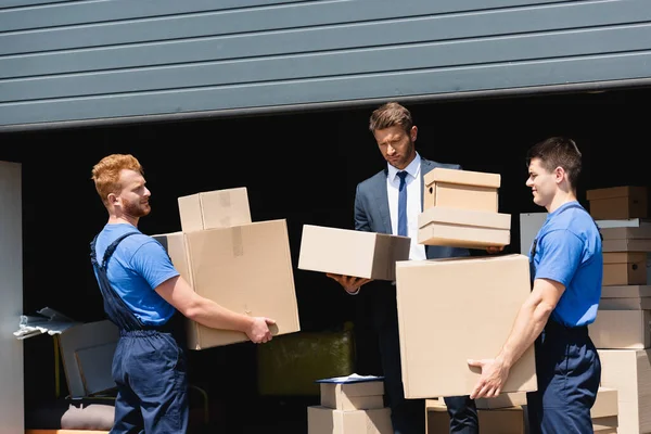 Confident businessman holding packages near movers and warehouse outdoors — Stock Photo