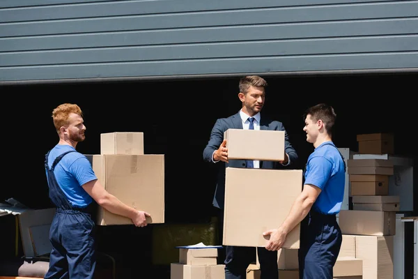 Homem de negócios em pacotes formais de empilhamento de desgaste perto de movers que trabalham ao ar livre — Fotografia de Stock