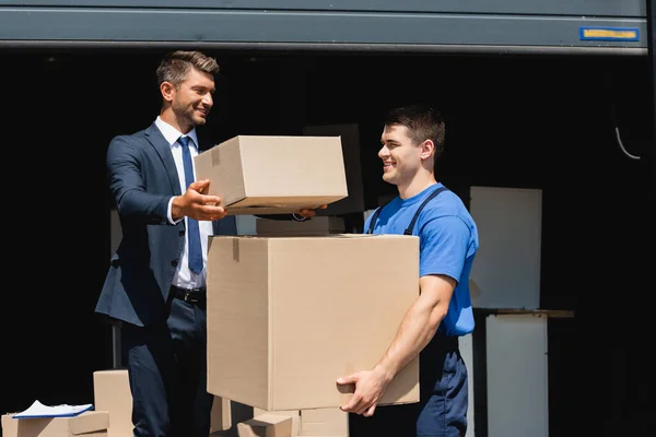 Homem segurando pacote perto de carregador com caixa de papelão e armazém ao ar livre — Fotografia de Stock