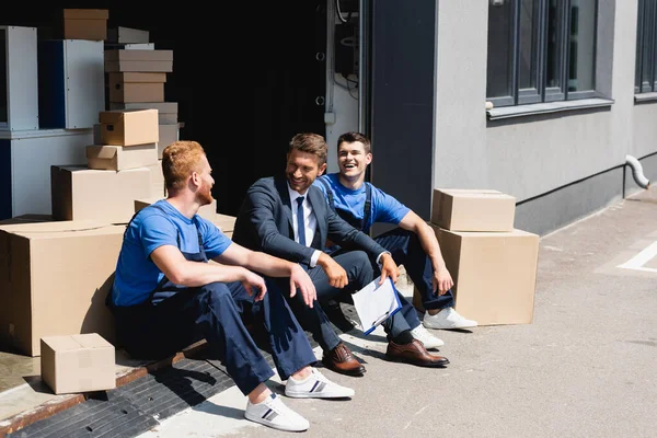 Homme d'affaires avec presse-papiers riant avec chargeuses près de boîtes en carton dans un entrepôt à l'extérieur — Photo de stock