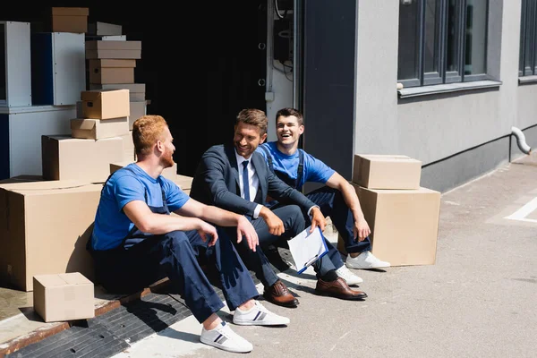 Businessman with clipboard laughing with loaders near cardboard boxes in warehouse outdoors — Stock Photo