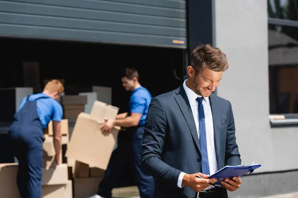 Selective focus of manger looking at clipboard while loaders working in warehouse outdoors — Stock Photo
