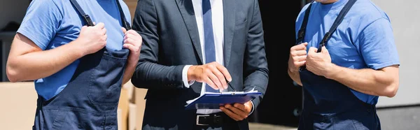 Panoramic crop of businessman with pen and clipboard near movers in overalls outdoors — Stock Photo