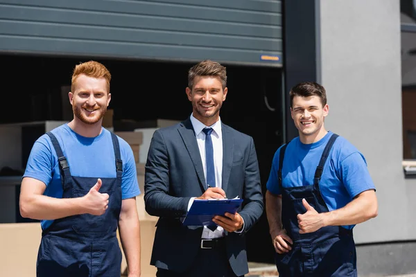 Movers showing thumbs up at camera near businessman writing on clipboard outdoors — Stock Photo