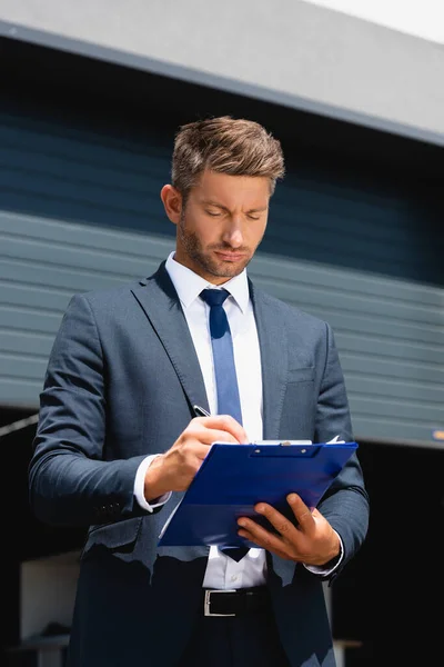 Businessman in suit writing on clipboard with warehouse on background — Stock Photo