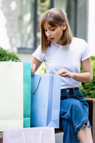 Selective focus of excited woman looking at shopping bag on bench on urban street — Stock Photo