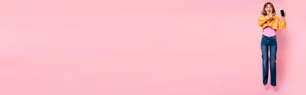 Panorama-Ausschnitt einer aufgeregten Frau, die mit dem Finger auf ihr Smartphone zeigt und auf rosa Hintergrund springt — Stockfoto