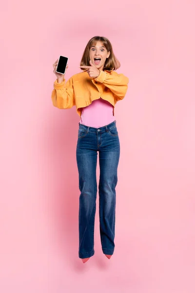 Excited woman jumping and pointing with finger at smartphone on pink background — Stock Photo