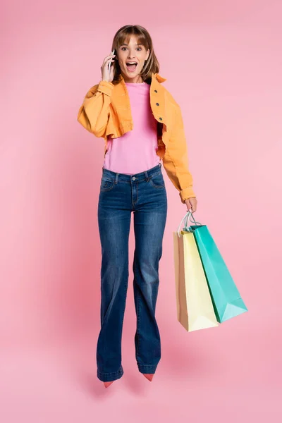 Захоплена жінка тримає сумки для покупок і розмовляє на смартфоні, стрибаючи на рожевому фоні — стокове фото