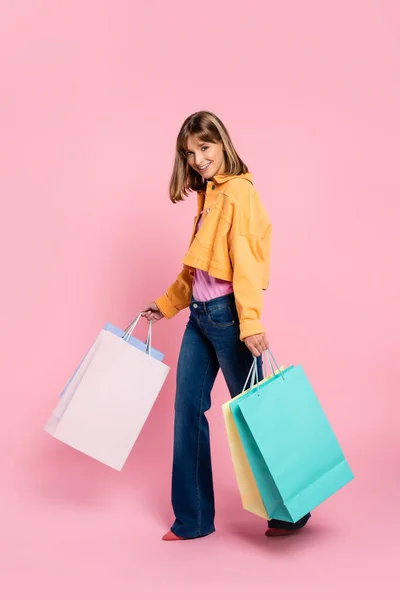 Femme en veste jaune regardant la caméra tout en tenant des sacs à provisions sur fond rose — Photo de stock