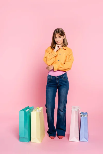 Jovem mulher olhando para a câmera perto de sacos de compras coloridos no fundo rosa — Fotografia de Stock