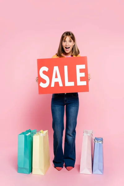 Emocionado joven mujer celebración de la tarjeta con la venta de letras, mientras que de pie cerca de bolsas de compras sobre fondo rosa - foto de stock