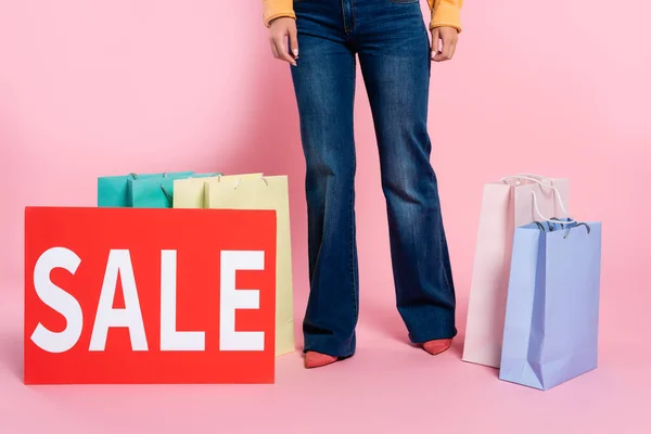 Vista recortada de la mujer de pie cerca de la tarjeta con letras de venta y bolsas de compras de colores sobre fondo rosa - foto de stock